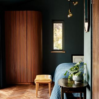 A bedroom with dark green wall paint decor, bench at end of bed, bed with blue throw and dark brown wooden table with indoor houseplant in planter