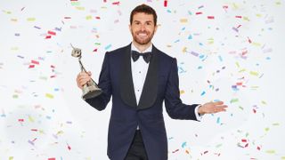  Host Joel Dommett in a suit and bowtie, holding an award and surrounded by falling confetti