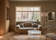 cozy living room with roman blinds, curved mohair sofa, round jute rug on light timber floor, glass coffee table, silver bar cart, floor lamp, artwork, white and chrome armchair and built-in timber bookcase, and sliding doors leading to the next room
