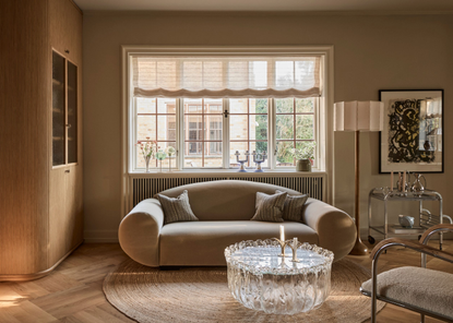 cozy living room with roman blinds, curved mohair sofa, round jute rug on light timber floor, glass coffee table, silver bar cart, floor lamp, artwork, white and chrome armchair and built-in timber bookcase, and sliding doors leading to the next room