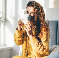 Woman in yellow pajama sitting on bed and drinking coffee at the sunny morning bedroom. - stock photo