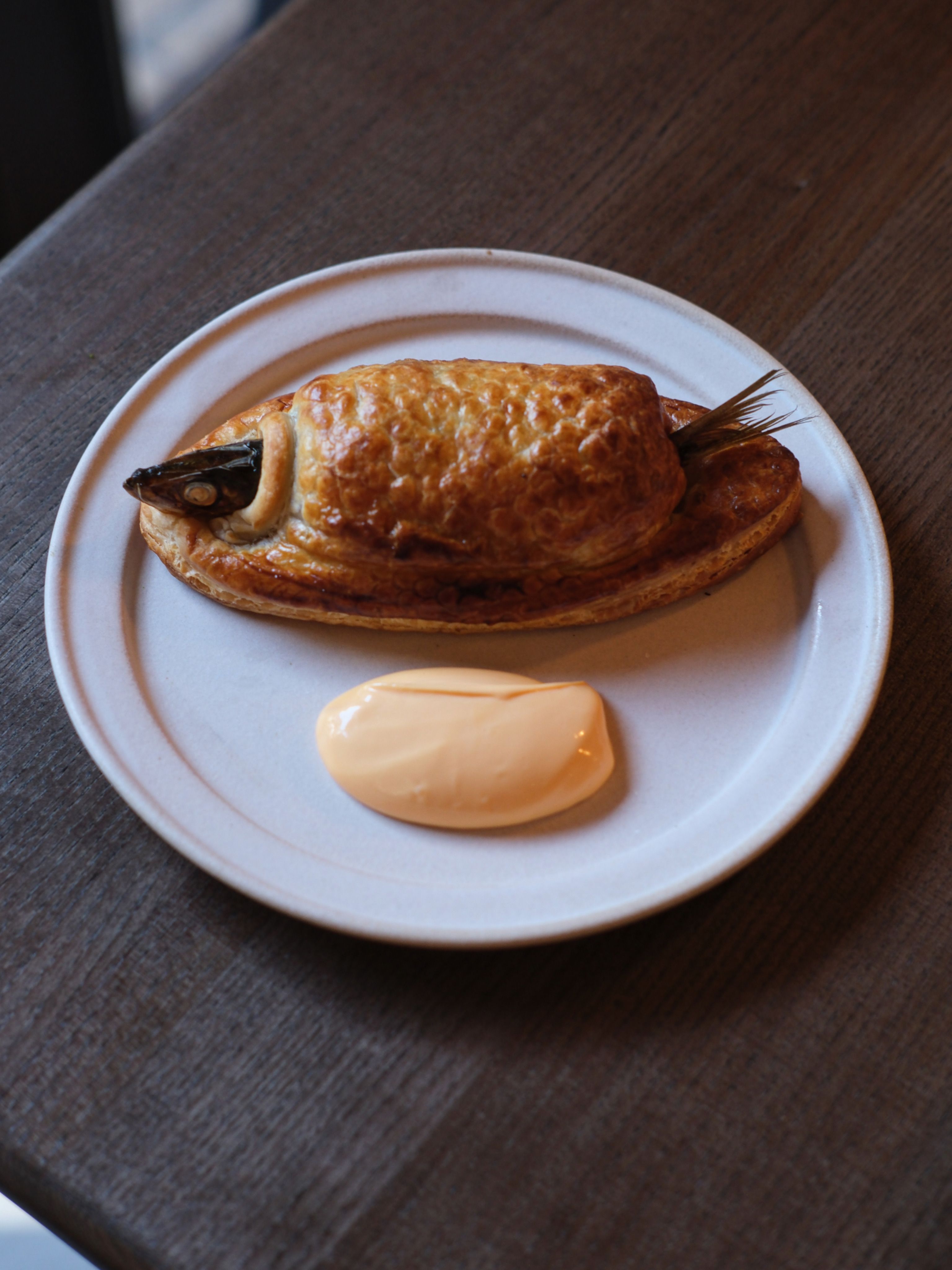 An en-croute fish fillet sits in a ceramic plate placed atop a wooden table next to a orange-looking mayo dip.