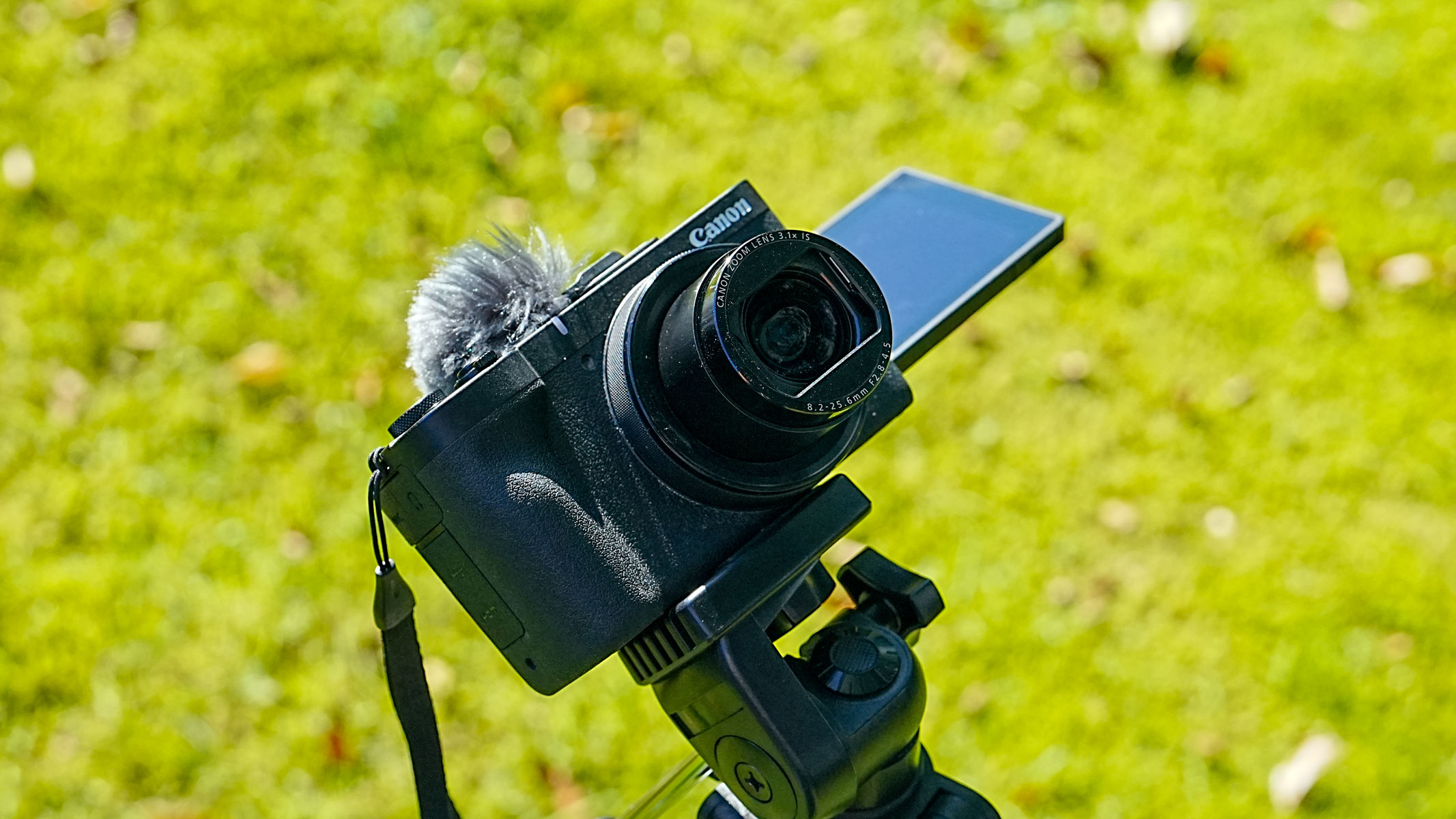 A Canon Powershot V1 with shutter open on a tripod with grass in the background.