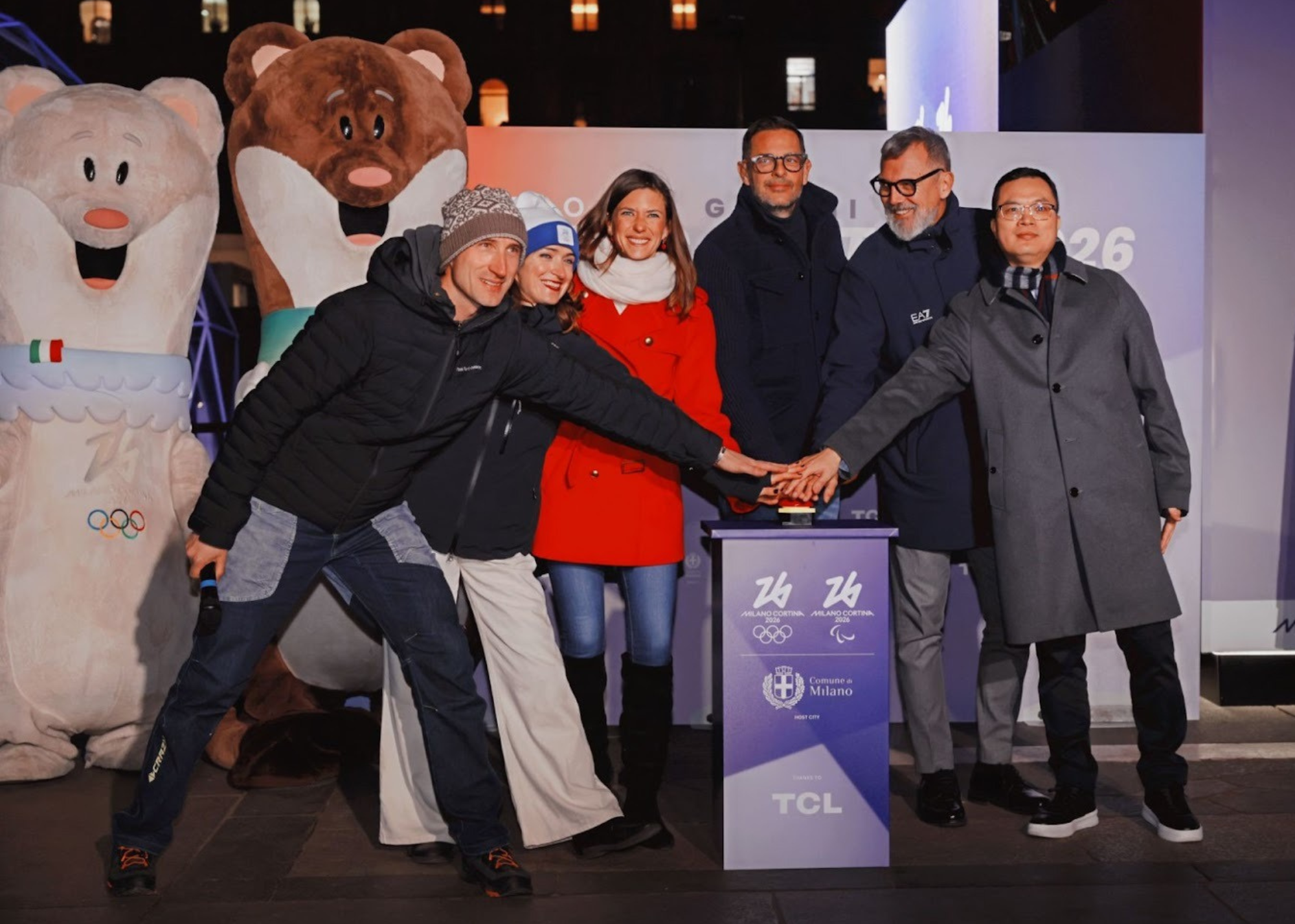 Five people pressing a red buzzer on a blue pedestal by mascots for the winter Olympics