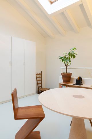 Interior view of a space on the upper floor of the Formafantasma Milan studio featuring light blue flooring, light coloured walls, a round table, a 'ZigZag' chair, a tall white storage unit, a wooden chair and a green plant in a pot on top of a wooden shelving unit
