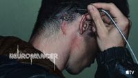 a person holds a wire with a male jack on the end of it up to their head, where a female plug can be seen surgically embedded into their skull behind their ear