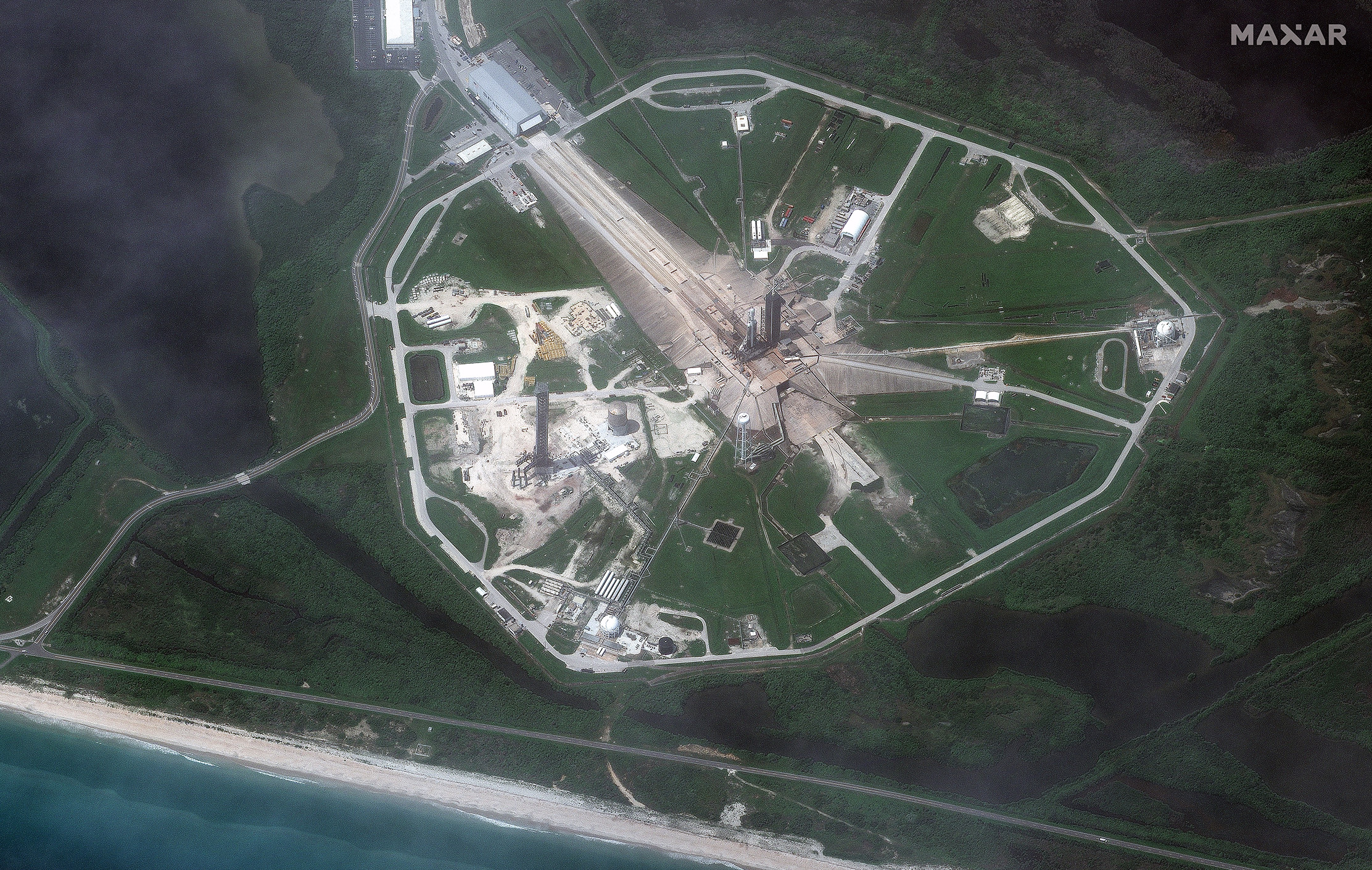 A high-altitude aerial view of SpaceX's triple-booster Falcon Heavy rocket can be seen standing in the center of the wide landscaped launch complex. Roads, pipes and other infrastructure extend outward from the rocket and adjacent launch tower.