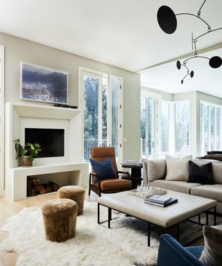 Living room space with tall floor to ceiling windows, painted gray-beige walls, white painted ceiling, modern rectangular fireplace design, sheepskin rug, brown leather lounge chair and two brown footstools, large square leather footstool, black hanging geometric mobile, gray sofa