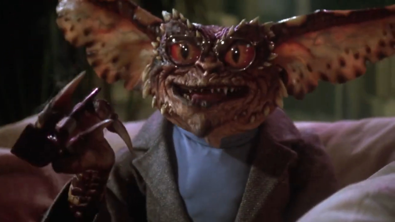 Brain Gremlin se sienta sosteniendo una pipa mientras está vestido con una chaqueta y un jersey de cuello alto en Gremlins 2: The New Batch.