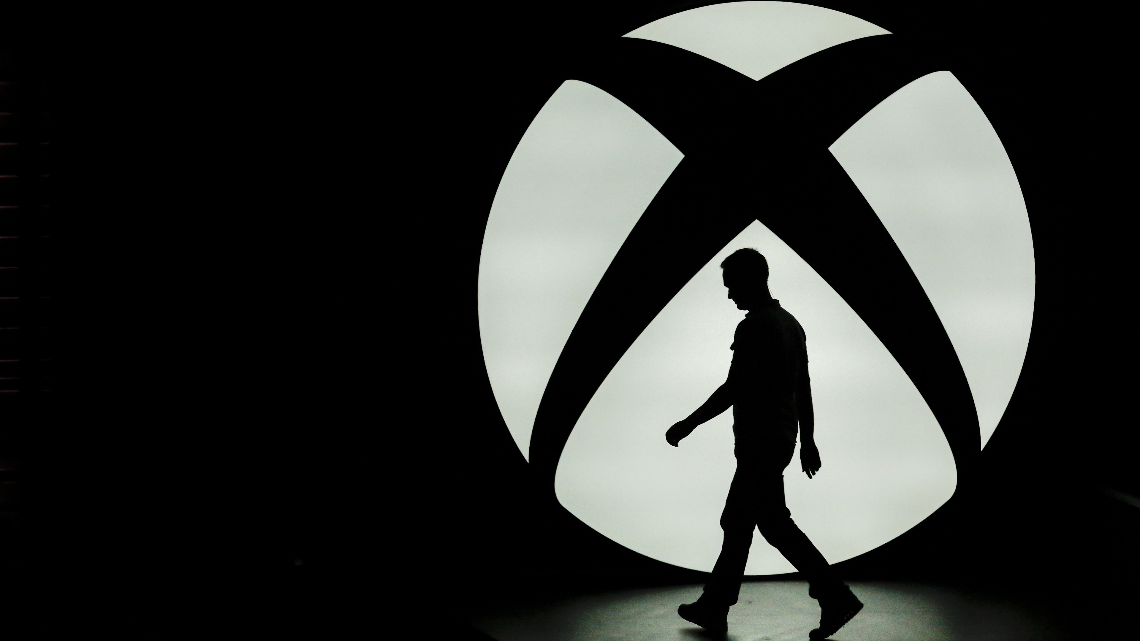 The silhouette of Ralph Fulton, design director for Forza Horizon at Playground Games, is seen walking past the Microsoft Corp. Xbox One logo uring a media event ahead of the E3 Electronic Entertainment Expo in Los Angeles, California, U.S., on Monday, June 9, 2014. Microsoft Corp. and Sony Corp. are gearing up for the next phase of competition between their dueling video-game consoles, highlighting exclusive titles that will be on sale for the year-end holiday season. Photographer: Patrick T. Fallon/Bloomberg via Getty Images 