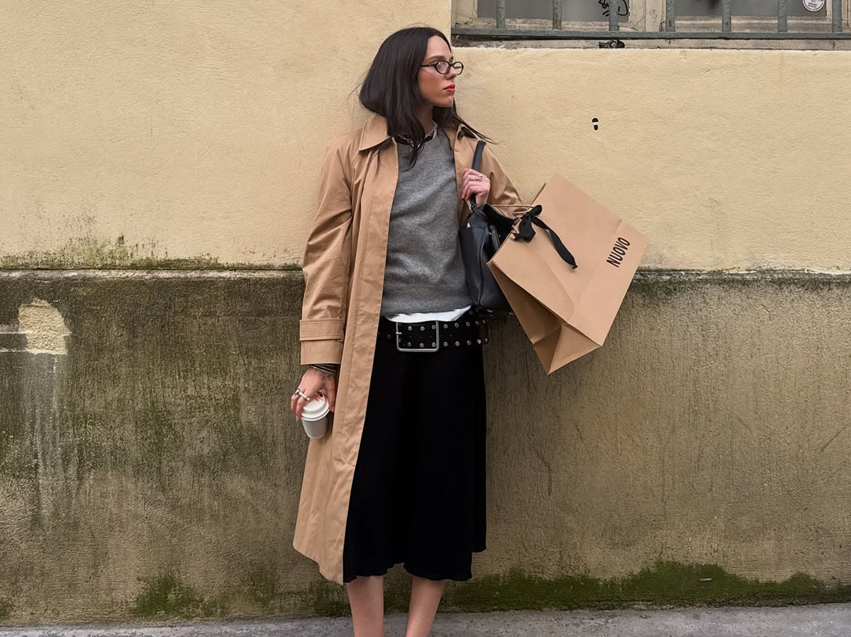 A woman wearing a tan trench coat, black culottes, white flats, and a gray sweater