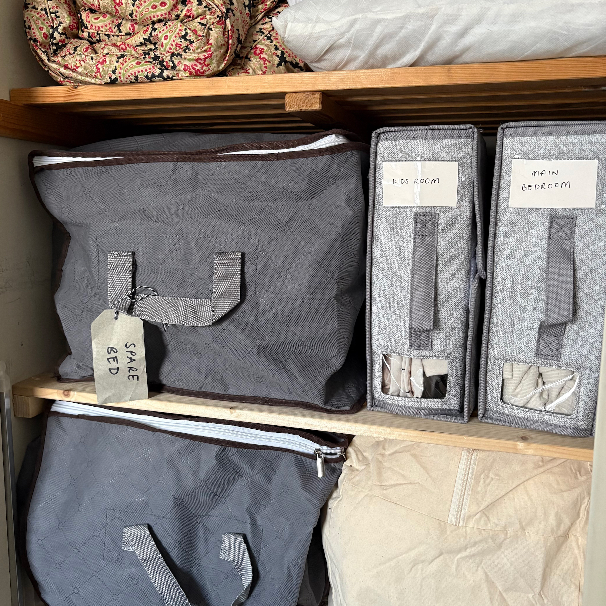 Two grey bedding organiser files next to a large grey clothes storage bag on a wooden shelf in an airing cupboard