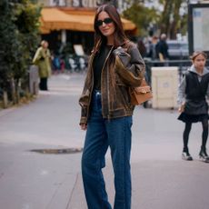 A woman wears a brown leather jacket, jeans, a black tee, and a tan leather bag.