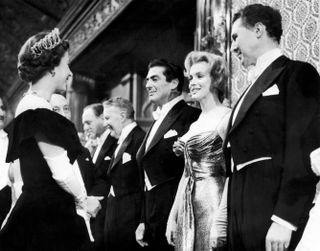 Queen Elizabeth wearing a tiara and gown talking to Marilyn Monroe wearing an evening gown and standing next to men in tuxedos