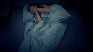 photo of a woman in with long, dark hair lying in bed under a large blanket. She holds both hands over her face as if struggling to sleep