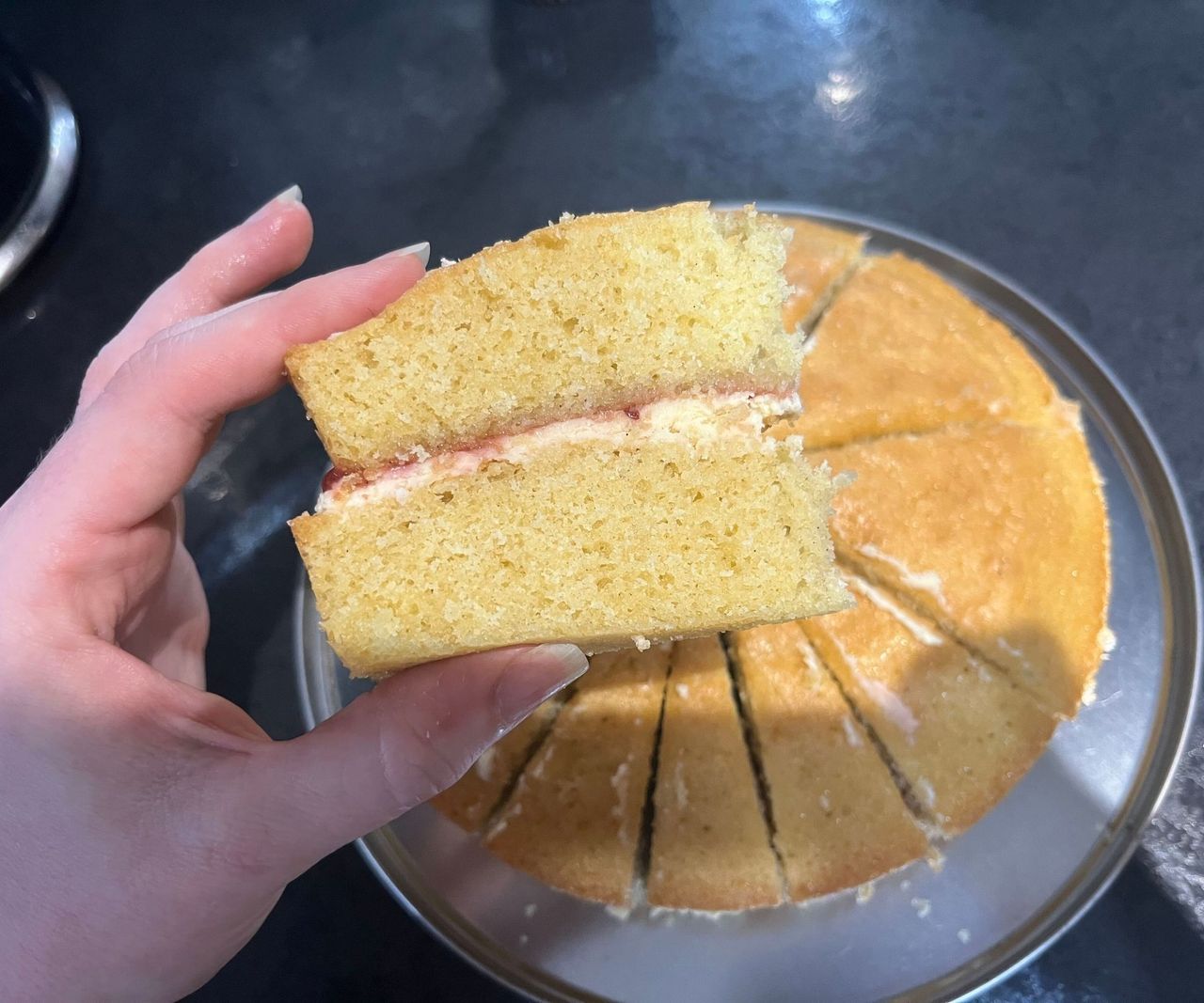 A piece of Victoria sponge cake being held above a whole cake on a black kitchen counter