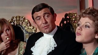 George Lazenby speaking while flanked by two women in On Her Majesty's Secret Service. 