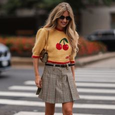 Emili Sindlev at New York fashion week wearing a cherry wool jumper and plaid skirt as she looks down smiling as she crosses the street in black sunglasses