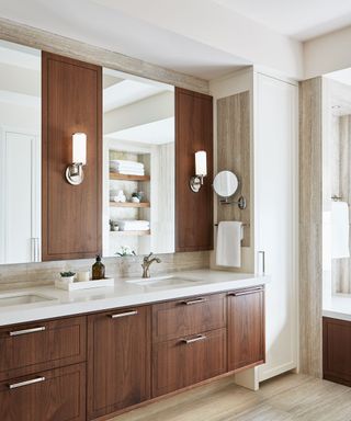 A double vanity bathroom with walnut colored walls and drawers, two mirrors and silver wall sconces on the wall, a white sink surface with a soap dispenser and plant, and stone colored wooden panels and flooring