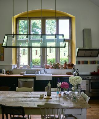 white kitchen with yellow paint inside of window, farmhouse style kitchen, pendant light, blue cabinets, flowers in vases on table, chairs