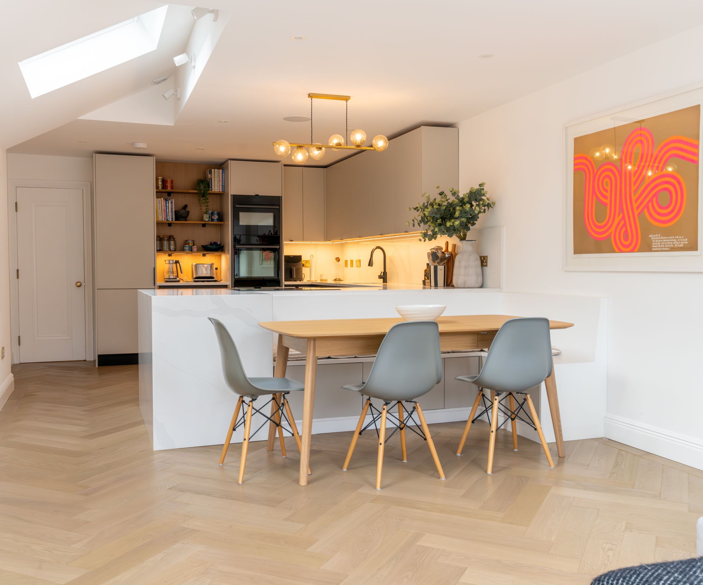 A kitchen with white walls and a wooden herringbone floor and electric orange wall art