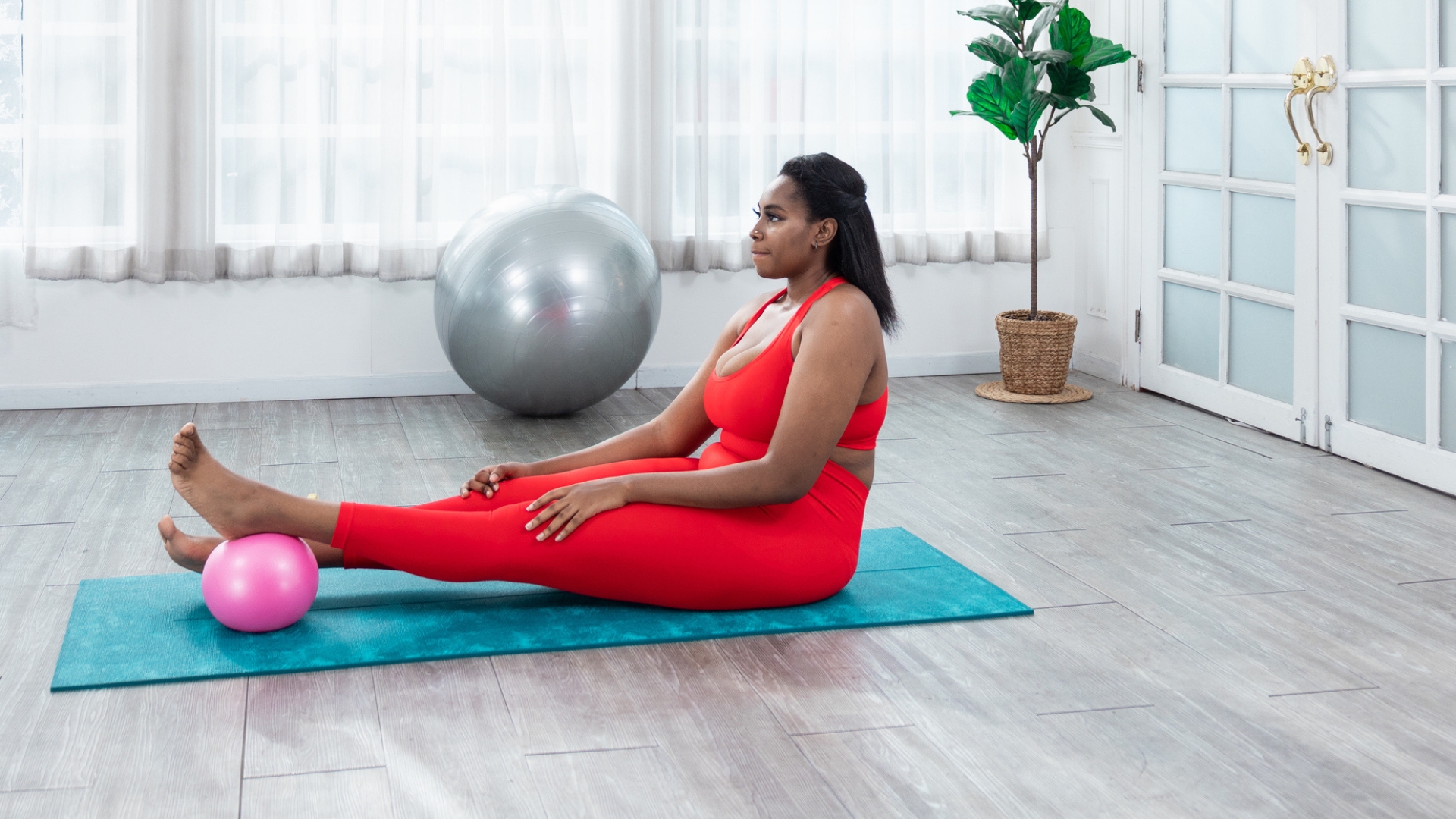 woman wearing red leggings and crop top sitting with legs outstretched on a turquoise exercise mat. there&#039;s a pink pilates ball under one foot and she&#039;s in a studio setting.