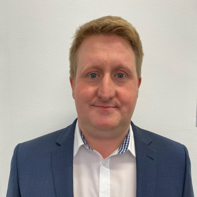 Headshot of a blonde man smiling at the camera wearing a blue suit and white shirt with a blue and white chequered pattern on the collar 