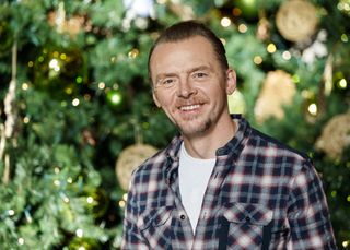Simon Pegg stands in front of an out-of-focus green backdrop of lit-up Christmas trees, wearing a navy, red and blue checked shirt over a white t-shirt and smiling