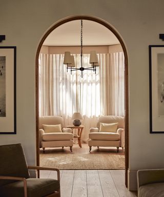 neutral living room with two armchairs and sheer curtains with an archway into the next room