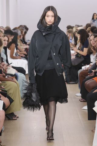 An SS24 runway photo at Proenza Schouler that shows a black nylon sporty jacket with a black midi skirt, tights, and thong sandals.