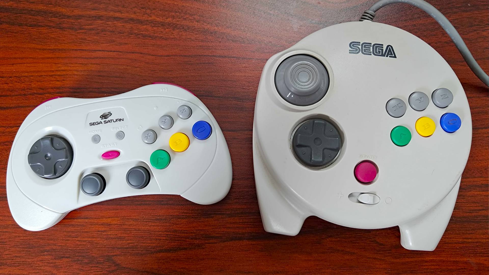 Retro-Bit Sega Saturn Pro controller on left and 3D Control Pad on right on woodgrain desk.