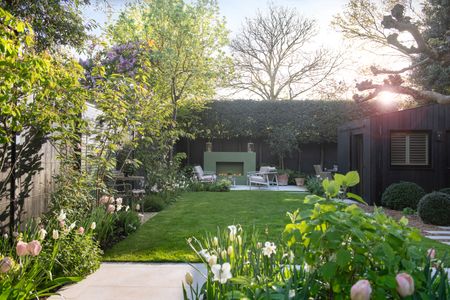 a garden with a large lawn, and multiple seating areas, with a fireplace, and large fence with hedge at the rear