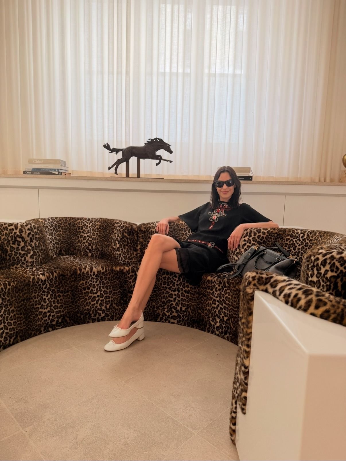 Alexa Chung sitting on a leopard-print couch wearing a black knee-length dress with white ballet pumps.
