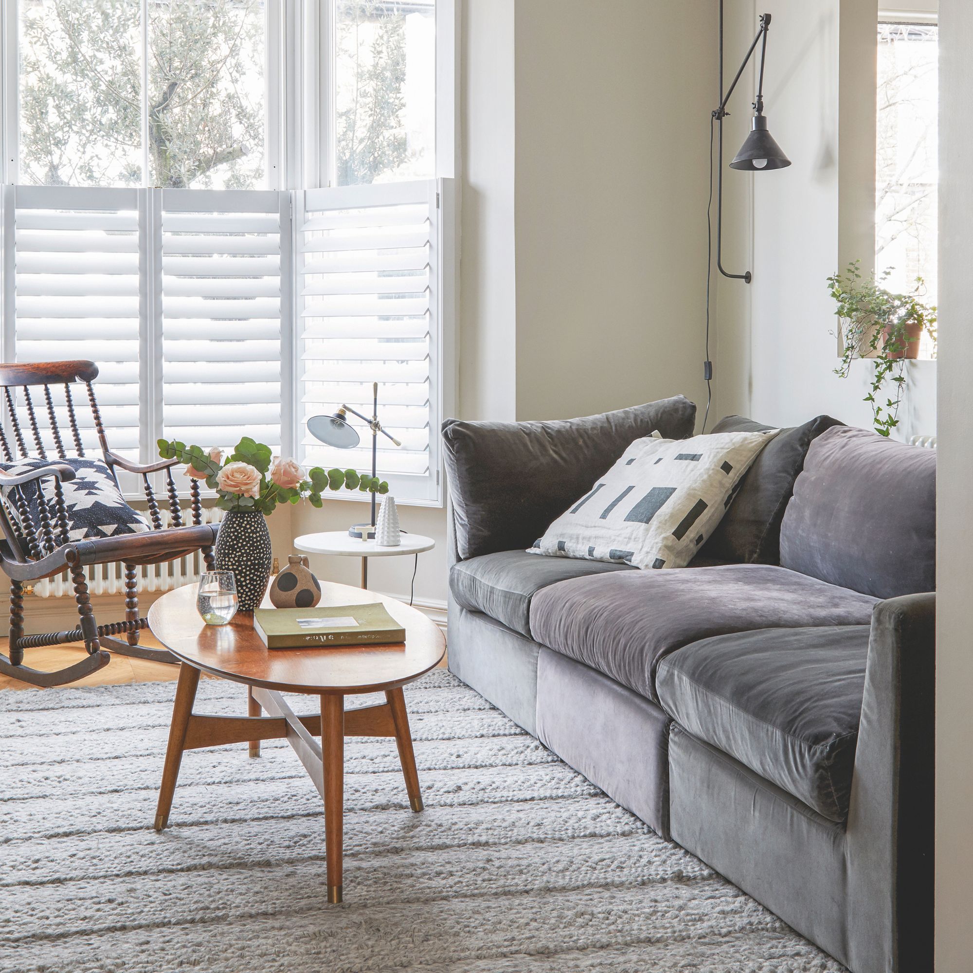 Plush grey sofa in a living room, with wooden coffee table and rocking chair nearby and open half-height shutters on the bay window
