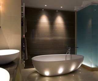 freestanding white bath with small LED lights in floor around the bath and blue glass wall lit from the base to the one side