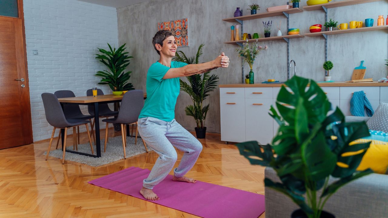 woman doing a half squat in a living room setting on a pink exercise mat. she&#039;s wearing a turquoise tshirt and there are colourful crockery items on kitchen shelves behind her.