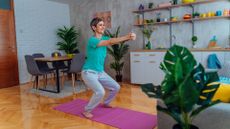 woman doing a half squat in a living room setting on a pink exercise mat. she's wearing a turquoise tshirt and there are colourful crockery items on kitchen shelves behind her.