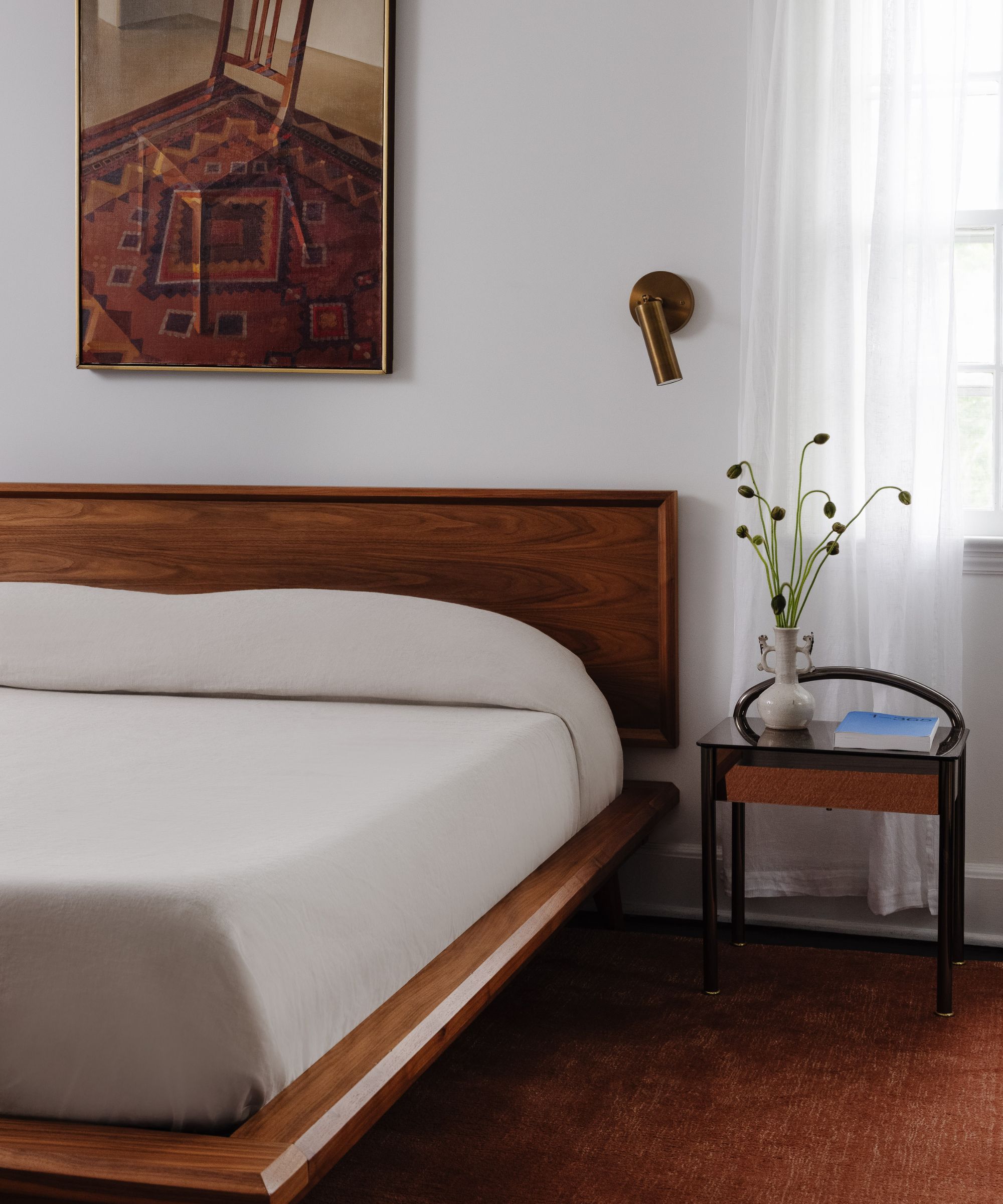 Bedroom with a wooden midcentury low bed, neutral linen bedding, a gold wall sconce, a still life painting of a chair, a dark red rug, and a wooden nightstand with a vase of flowers.