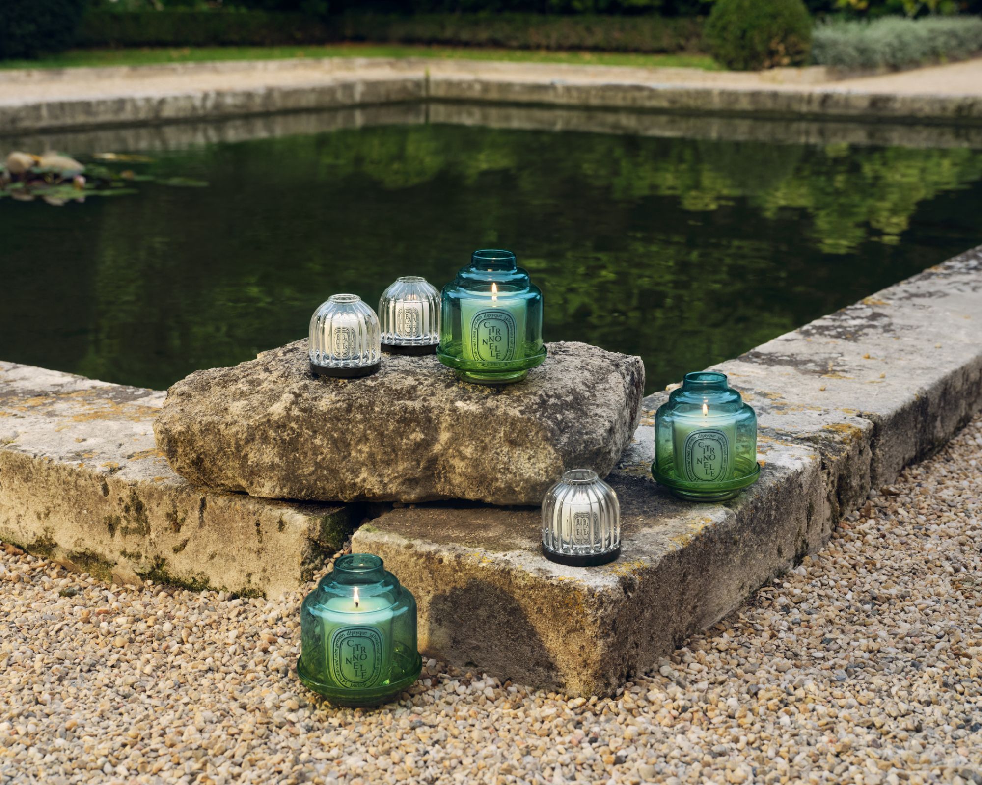 Diptyque citronella collection pictured on a gravel and stone surface surrounding a large square pond.