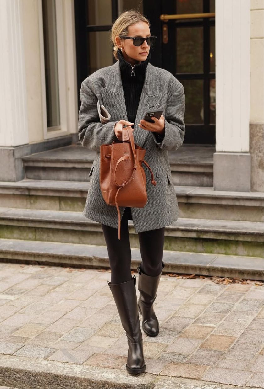 @anoukyve wears a grey wool blazer, black sweatshirt, brown leggings, riding boots and a tan leather bucket bag