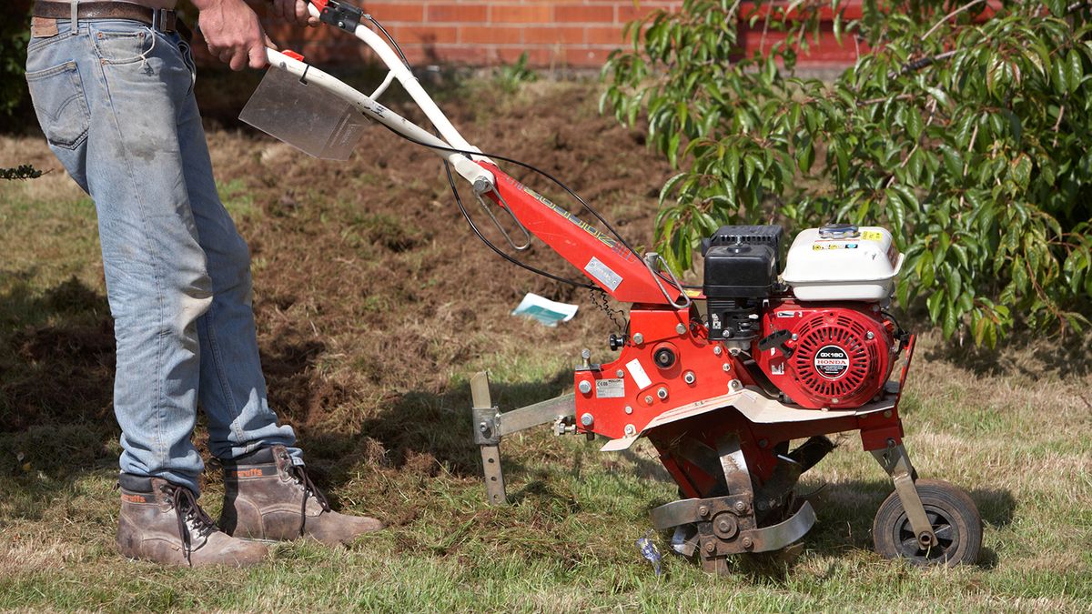 man wearing jeans and safety boots operates petrol driven garden rotavator being used to churn up soil in a garden