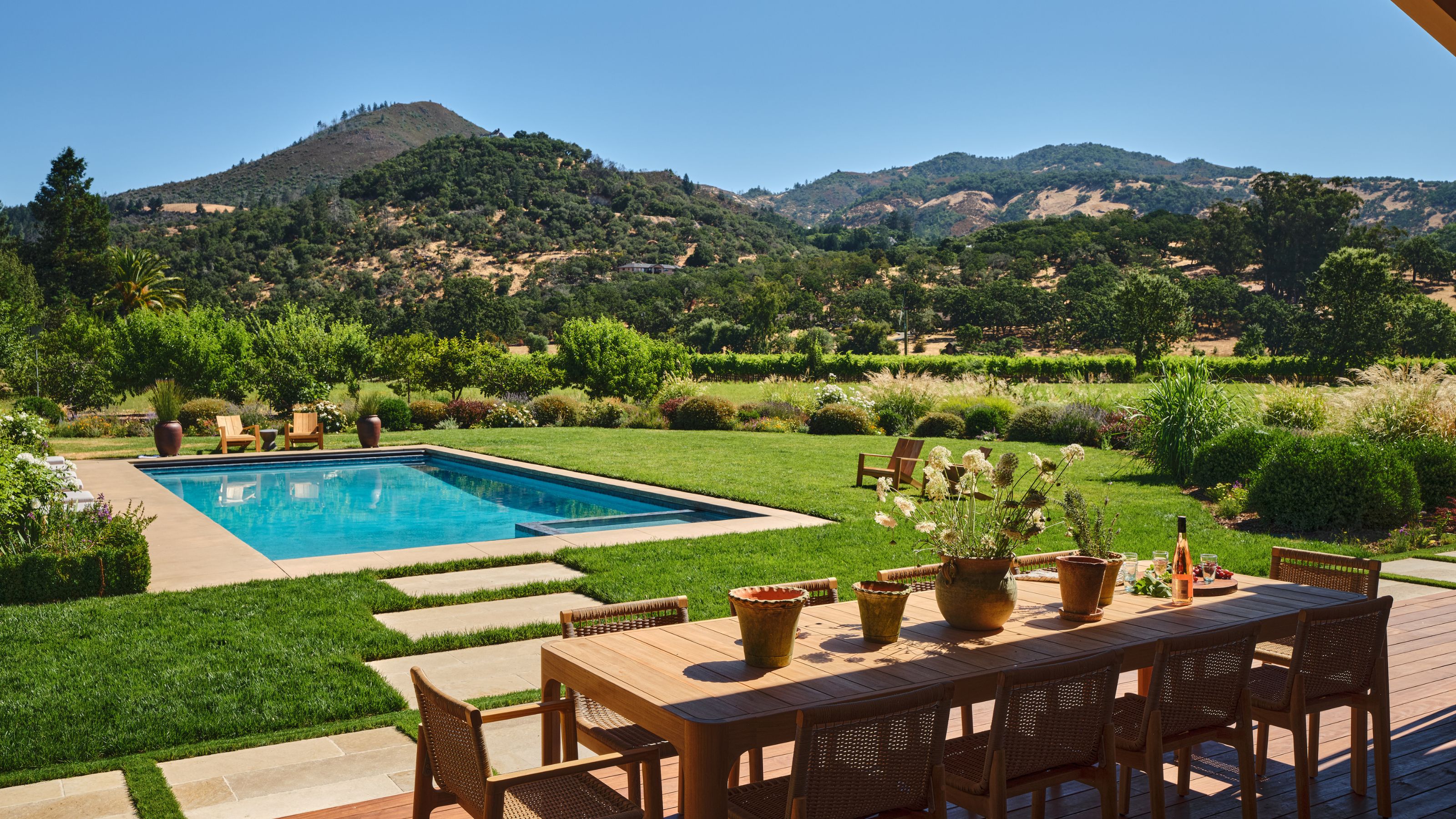 a deck area set up with a wooden dining set overlooking a large pool and a view to rolling hills and vineyards