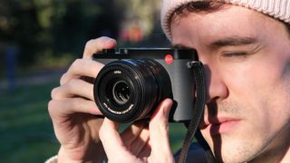 A person holding a Leica Q3 43 camera up to their face in bright sunlight