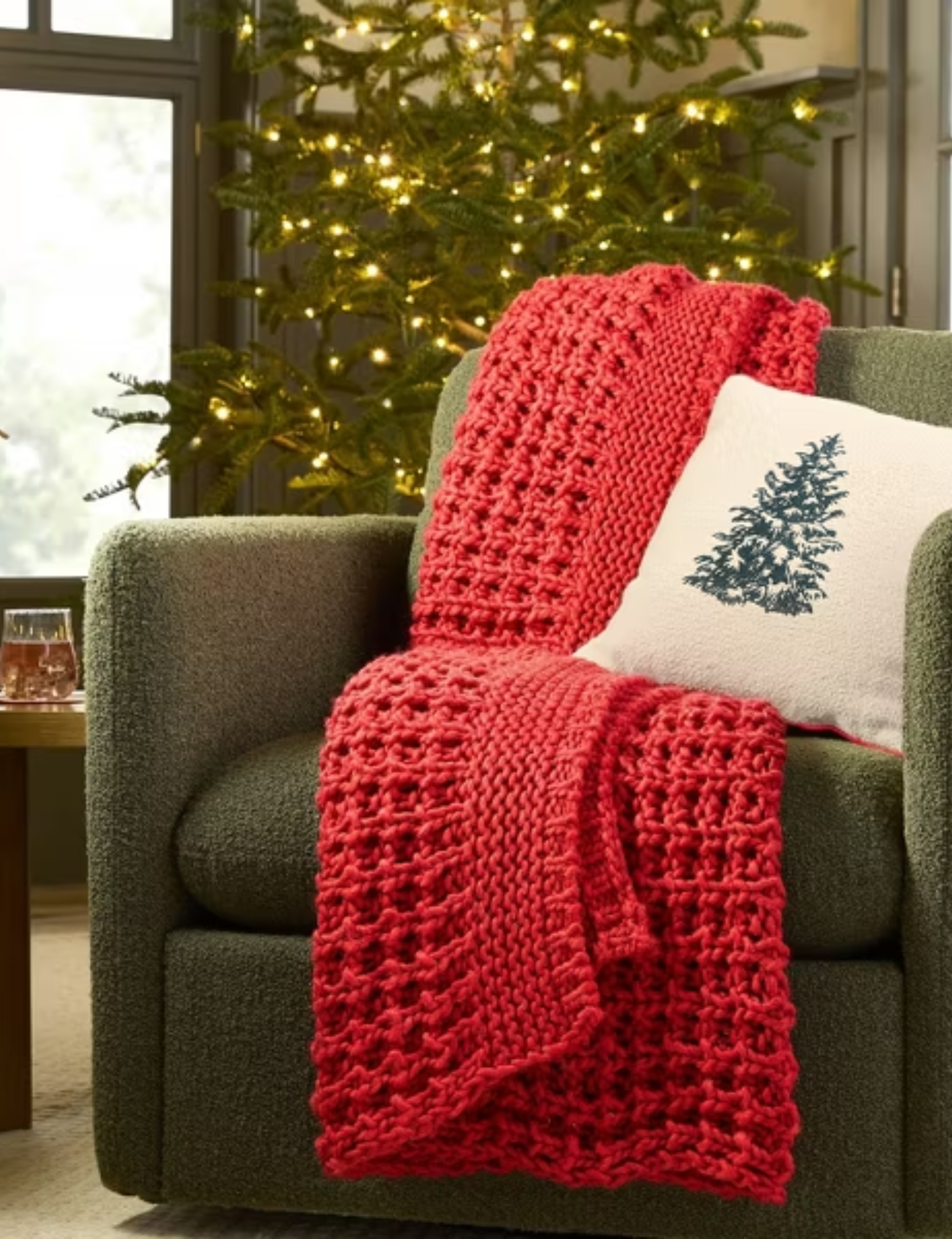 Red chunky knit throw blanket and Christmas-tree motif throw pillow draped overtop of an evergreen accent chair