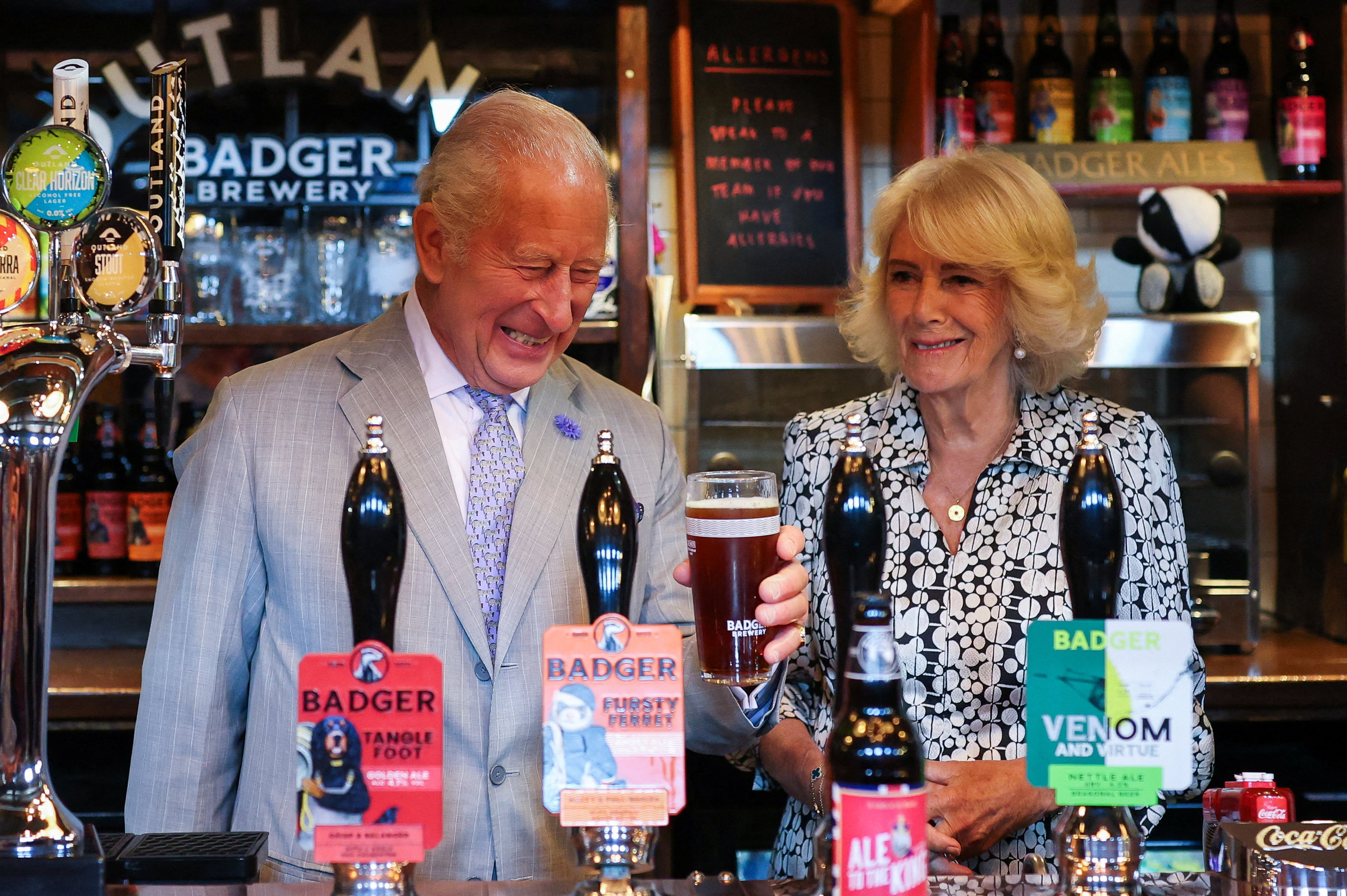 Queen Camilla and King Charles laughing while pulling pints at a pub