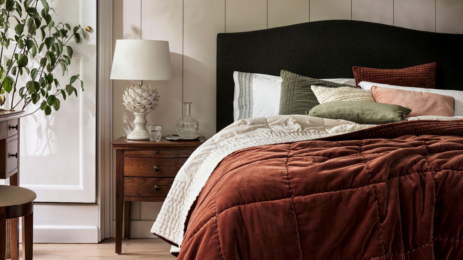 A bed with a red velvet throw on it, and matching red and green pillows. A small antique-style wooden nightstand beside it, with a large artichoke-style lamp with a fabric shade. 