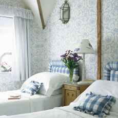 A blue and white delicately wallpapered twin bedroom with matching gingham headboards and cushions. 