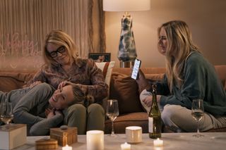 A woman (Kristen Bell as Joanne) lays her head on her mother's (Stephanie Faracy as Lynn) lap, as her sister (Justine Lupe as Morgan) shows them an iPhone message, all three on a living room couch, in episode 103 of Nobody Wants This.