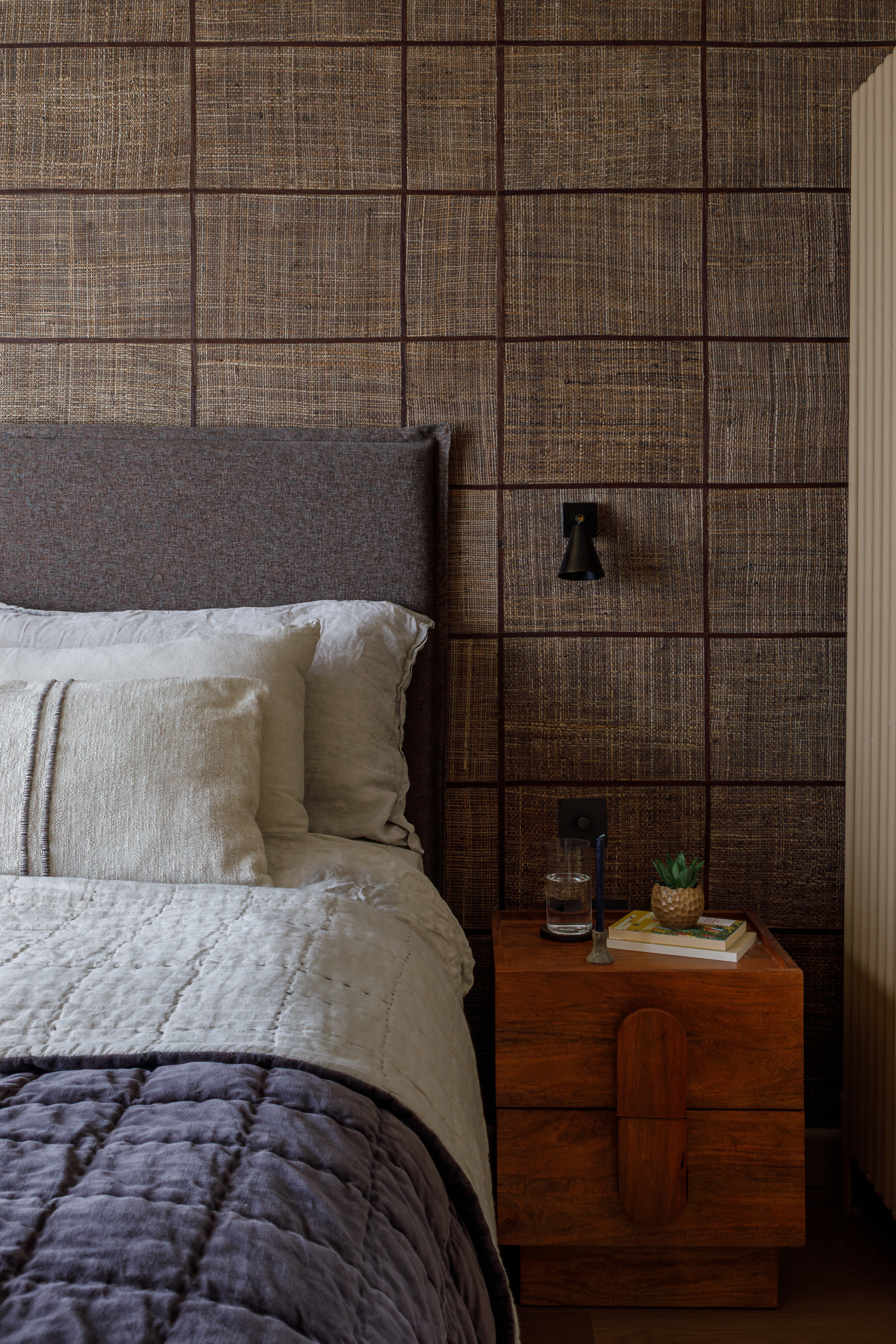bedroom with grasscloth wallpaper, timber side table, gray headboard, beige and navy linen sheets