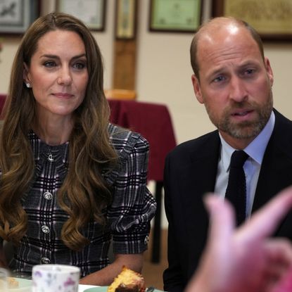 Kate Middleton wears a checked coat and has long wavy brunette hair while sitting next to Prince William